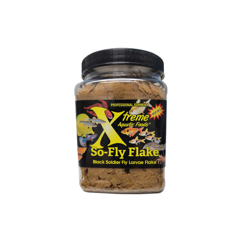 A clear plastic jar containing Xtreme 99g So-Fly Flake fish food, with a black lid and a label featuring the product name and branding with various different tropical fish.